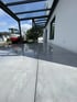 Moderne Terrasse mit grauen großformatigen Fliesen unter einer Glasüberdachung mit Metallrahmen neben einem weißen Haus.
