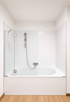 Weiße Badewanne mit gläserner Duschwand, Handbrause und hellen Metro-Fliesen in einem Badezimmer mit Holzboden.