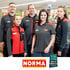 Fünf lächelnde NORMA-Mitarbeiter in roten und grauen Uniformen stehen in einem Supermarktgang.