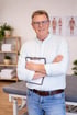 Ein lächelnder Physiotherapeut mit Brille und Klemmbrett steht vor einer Behandlungsliege und anatomischen Postern.
