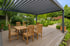 Holztisch und Stühle unter einer grauen Pergola auf einer Holzterrasse in einem Garten.