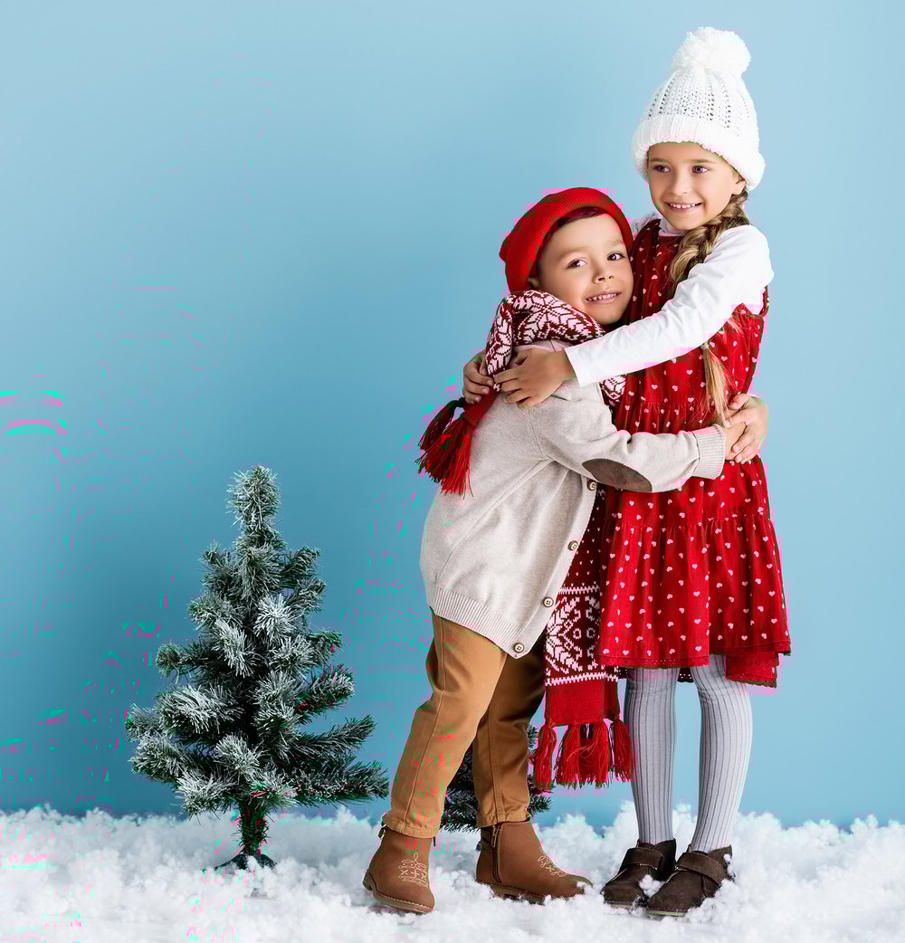 Zwei lächelnde Kinder in Winterkleidung umarmen sich vor blauem Hintergrund mit einem kleinen Weihnachtsbaum und Schnee.