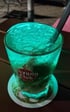 Glastasse mit leuchtend grünem Getränk, Eiswürfeln und einem schwarzen Strohhalm, mit "Havana Club" Logo.