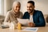 Lächelndes Paar schaut auf Dokumente mit Diagrammen; Modellhaus und Goldbarren auf dem Tisch.