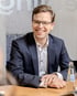 Ein lächelnder Mann in Anzug und Brille sitzt an einem Konferenztisch neben einer Frau.
