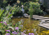 Teich mit Trittsteinen, umgeben von blühenden Pflanzen und Sträuchern, ein Gartenbrunnen im Hintergrund.