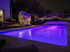 Ein beleuchteter Pool mit lila Licht im Garten bei Nacht.