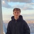 Lächelnder junger Mann mit lockigem Haar in einer schwarzen Kapuzenjacke vor einer Stadtkulisse bei bewölktem Himmel.