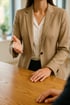 Eine Person in einem beigefarbenen Blazer und weißer Bluse gestikuliert über einem Holztisch.