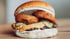 Ein Burger mit Zwiebelringen, Essiggurken und Soße auf einem Holzbrett.