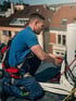 Techniker in blauer Kleidung und Sicherheitsgurt arbeitet an Kabeln auf einem Dach.