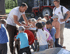 Ein Mann zeigt Kindern, wie man eine Milchzentrifuge bedient; im Hintergrund ein roter Traktor.