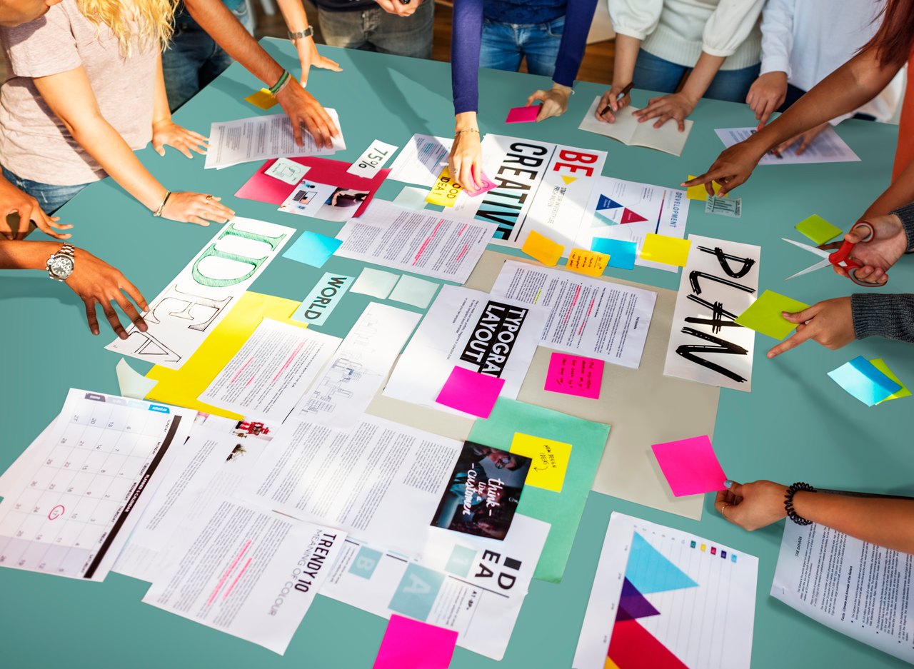 Mehrere Hände arrangieren Papiere, Notizzettel und Dokumente mit den Wörtern „BE CREATIVE“ und „PLAN“ auf einem Tisch.
