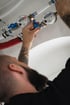 Ein Handwerker dreht an einem blauen Ventilhebel neben zwei Wasserzählern an weißen und roten Rohrleitungen.