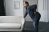 A man leans against a couch, holding his lower back.