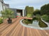 Ein Holzdeck umschließt einen Teich mit Seerosen und Schilf, daneben ein geöffneter Container und eine Palme.