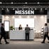 Messestand mit "MESSEN" Schild, Schalter, einer Mitarbeiterin und zwei Besuchern vor einem leeren Bildschirm.