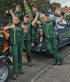 Fünf Landschaftsgärtner in grünen Uniformen lächeln und geben Daumen hoch vor einem Transporter und einem Gerät.