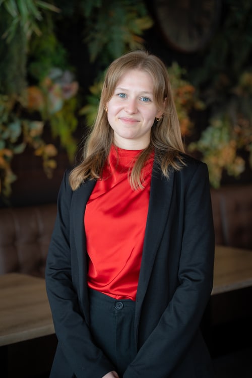 Junge Frau mit blonden Haaren, roter Bluse und schwarzem Blazer, lächelt in die Kamera.