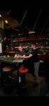 Zwei Männer in einer Bar, einer sitzt am Tisch und sieht aufs Handy, der andere filmt ihn.