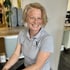 Eine lächelnde Frau mit blonden Haaren trägt ein graues Poloshirt mit einem weißen Logo.