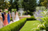 Brautpaar und Hochzeitsgäste gehen durch einen Garten mit Hecken und Springbrunnen.
