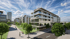 Moderne Mehrfamilienhäuser mit Glasbalkonen an einer gepflasterten Promenade mit Bäumen und Rasenflächen unter blauem Himmel.