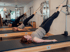 Drei Frauen trainieren in einem Studio an Pilates-Reformern mit in die Luft gestreckten Beinen.