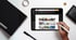 Eine Hand hält einen Stift und tippt auf ein Tablet, das eine Webseite mit Produktbildern anzeigt.