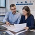 Zwei Kollegen besprechen technische Zeichnungen im Büro vor einer Wandgrafik zu Logistik und Produktion.