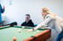 Lachende Frau spielt Billard, Mann sitzt lächelnd daneben, Kugel auf Billardtisch.