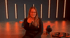 Frau in schwarzem Oberteil zeigt mit dem Finger, umgeben von orangefarbenen Fitnessgeräten und vertikalen Lichtern.