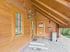 Eingangsbereich eines Blockhauses aus hellem Holz mit Sprossenfenster, überdachter Terrasse und gefliesten Stufen.