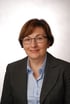 Frau mit braunem Bob, Brille, blauem Hemd und grauem Blazer lächelt.