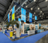 Messehalle mit TUI-Stand, blauer Teppich, Palmen, Bildschirme mit „GRECOTEL“ und „BLUE“ Logos.