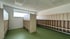 Leerer Umkleideraum mit Holzbänken und Regalen vor weißer Wand, grünem Boden und Deckenplatten.