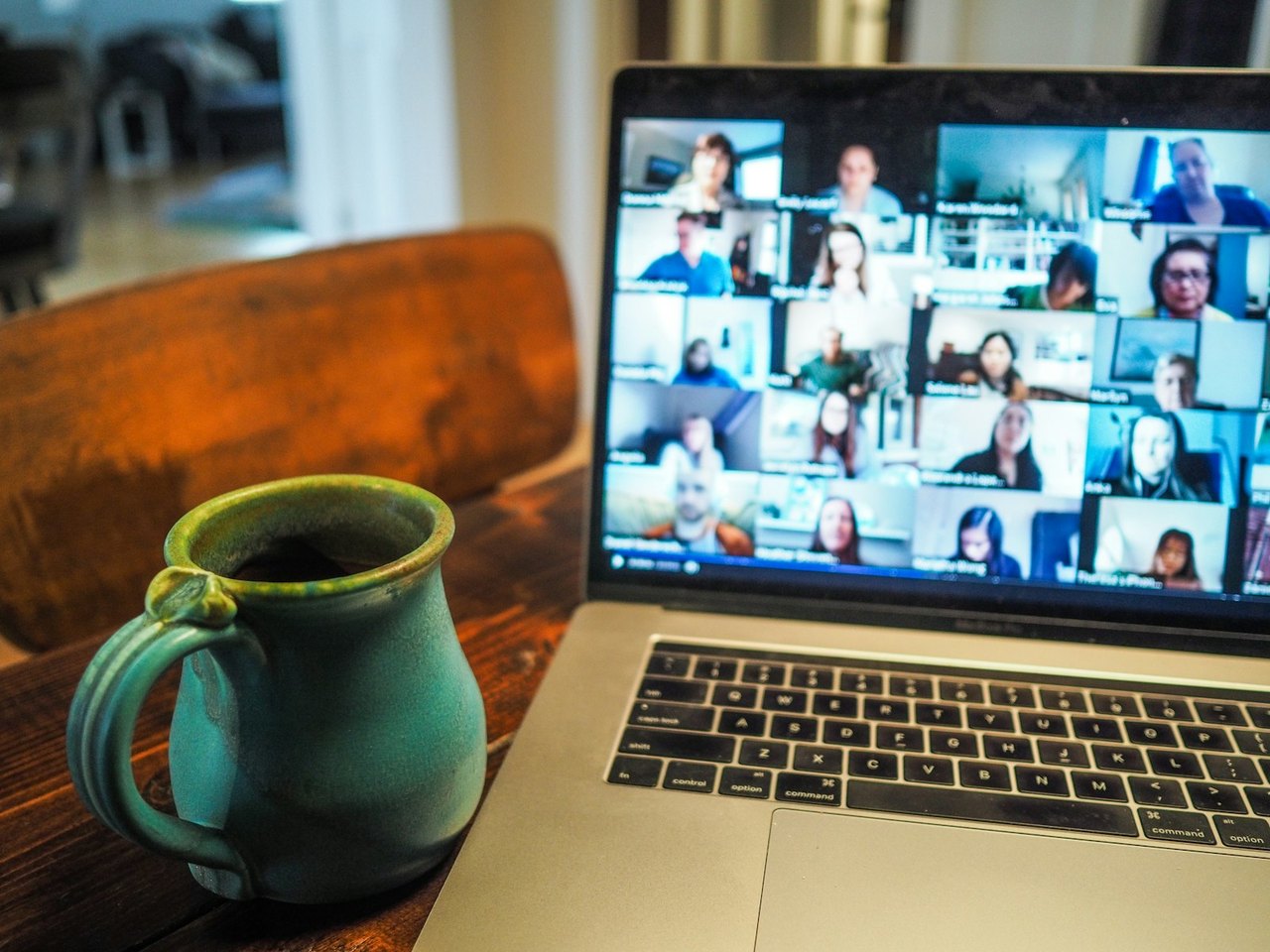 Aufgeklapptes MacBook auf einem Holztisch, auf dem Bildschirm eine Videokonferenz mit vielen Teilnehmenden, daneben ein grüner Keramikbecher