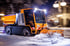 Oranger Schneepflug räumt nachts Schnee von einer Straße mit eingeschalteten Scheinwerfern.