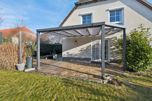 Graues Terrassendach mit weißer Stoffbespannung vor einem beigen Haus mit grüner Rasenfläche.