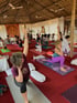 Menschen praktizieren Yoga-Übungen auf Matten in einem großen Raum mit Reetdach und Deckenventilatoren.