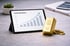 Tablet auf Schreibtisch mit Gold-Sparplan-Grafik, Goldbarren und Münzen im Vordergrund