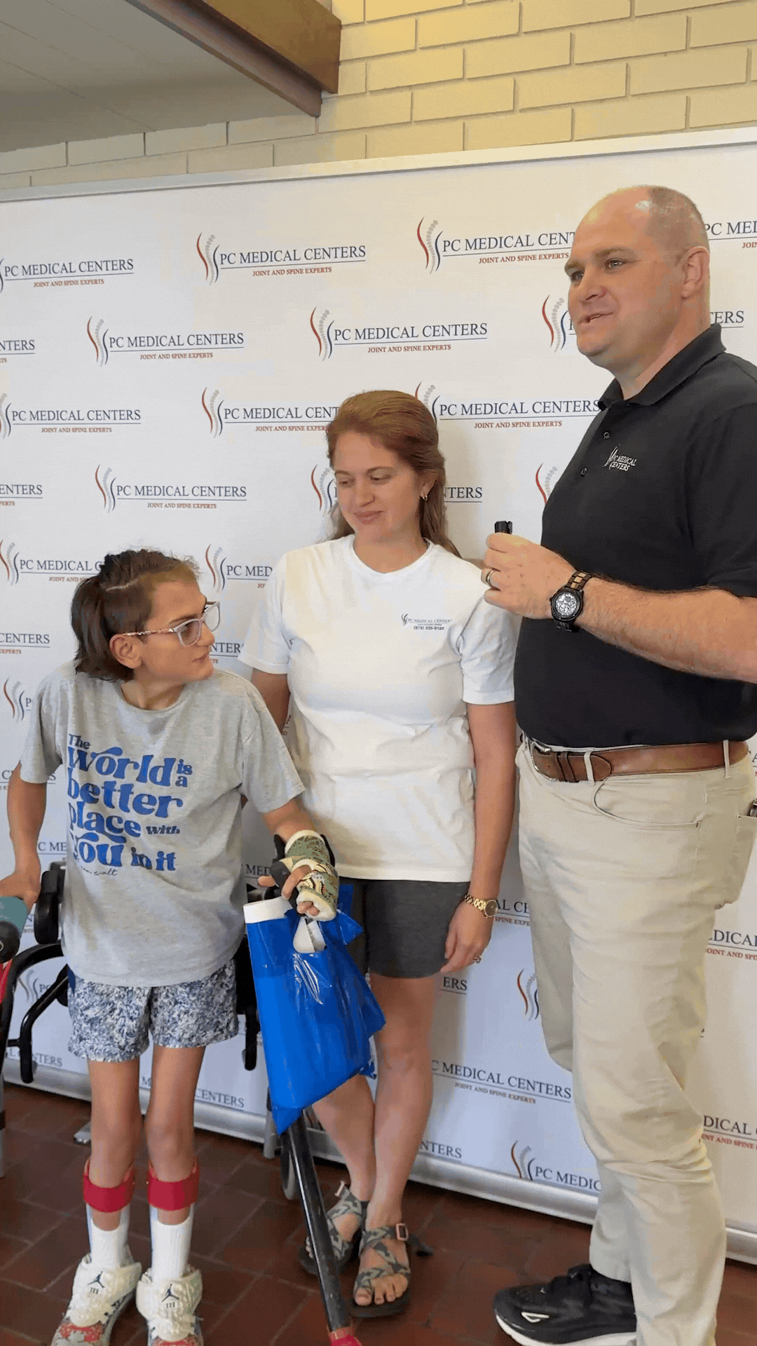 Young patient using a walker stands with a woman and a PC Medical Centers staff member in front of the clinic's backdrop