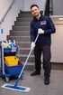 Ein lächelnder Mann in HSK-Arbeitskleidung wischt einen gefliesten Boden neben einem Putzwagen und einer Treppe.