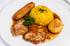 Gebratenes Fleisch mit gelbem Reis, einer Backbanane und einem Teigkloß in brauner Sauce auf einem weißen Teller.
