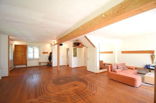 Großer, heller Raum mit Holzboden und Yin-Yang-Symbol, Holzbalken an der Decke und einer orangefarbenen Couch.