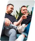 Zwei lächelnde Männer in schwarzen T-Shirts sitzen auf einem Sofa und blicken gemeinsam auf einen Laptop-Bildschirm.
