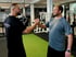 Zwei lächelnde Männer geben sich im Fitnessstudio die Hand. Der linke Mann trägt ein schwarzes T-Shirt mit der Aufschrift „DMITARENKO PERSONAL TRAINER“.