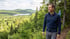 A man wearing a backpack and blue jacket hikes on a forest trail overlooking a lake and green mountains.