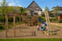 Ein Vater spielt mit seinem Kind auf einem großen Spielplatz mit Schaukeln und Klettergerüsten vor einem Holzhaus.