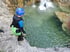 Ein Kind mit Helm und Neoprenanzug steht am Rand eines Wasserfalls, während eine Person ins Wasser springt.